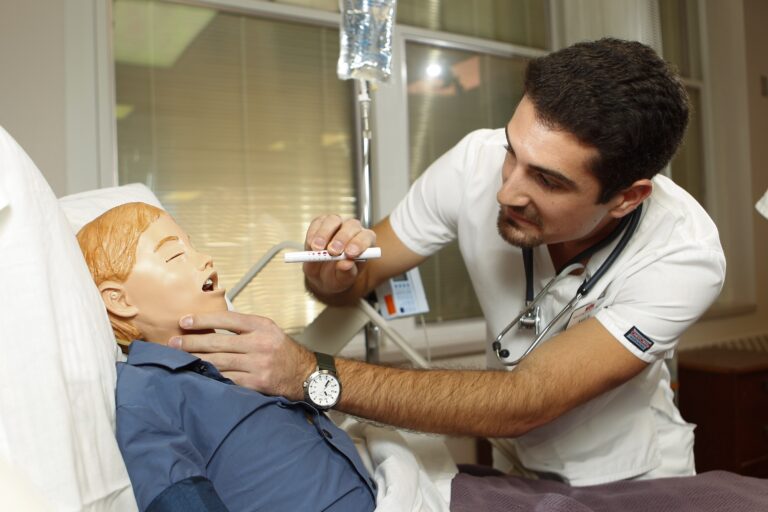 A male nursing student using a penlight to examine the throat of a pediatric mannequin in a hospital bed.