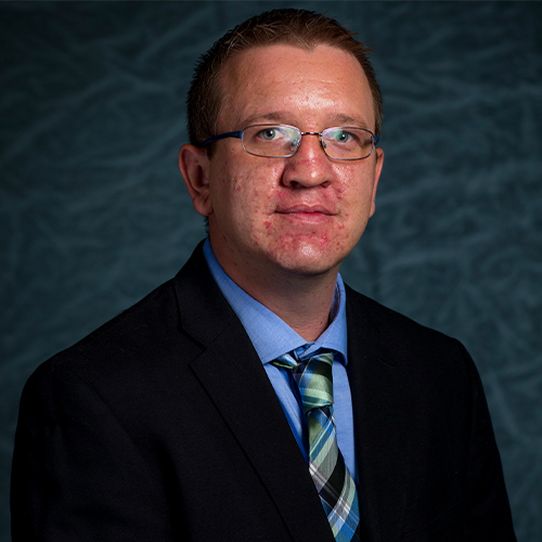 Headshot of Dr. Aaron Keller, Associate Professor of Chemistry at William Jewell College, wearing glasses, a suit jacket, and a plaid tie against a gray backdrop.