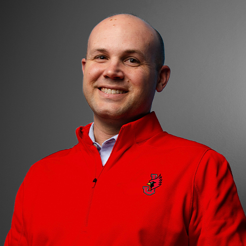 Ryan Erlacher, Vice President for Athletics at William Jewell College, smiling in a portrait while wearing a red Cardinal logo quarter-zip pullover.
