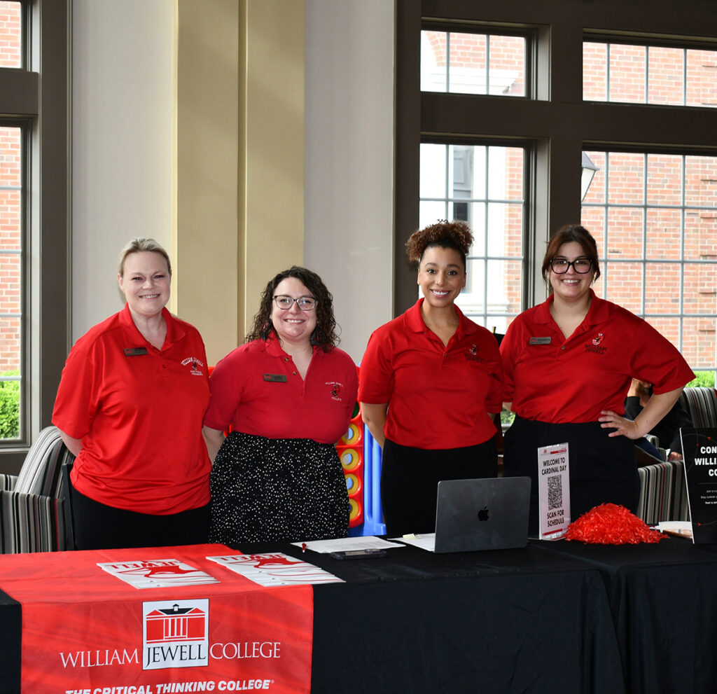 4 admission counselors wearing red William Jewell College polo shirts standing behind a welcome table during Cardinal Day.
