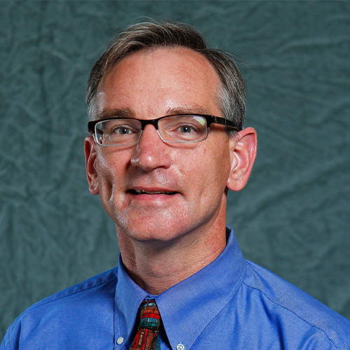 Headshot of Dr. Blane Baker, Professor of Physics at William Jewell College, wearing glasses, a blue button-down shirt, and a patterned tie against a gray backdrop.