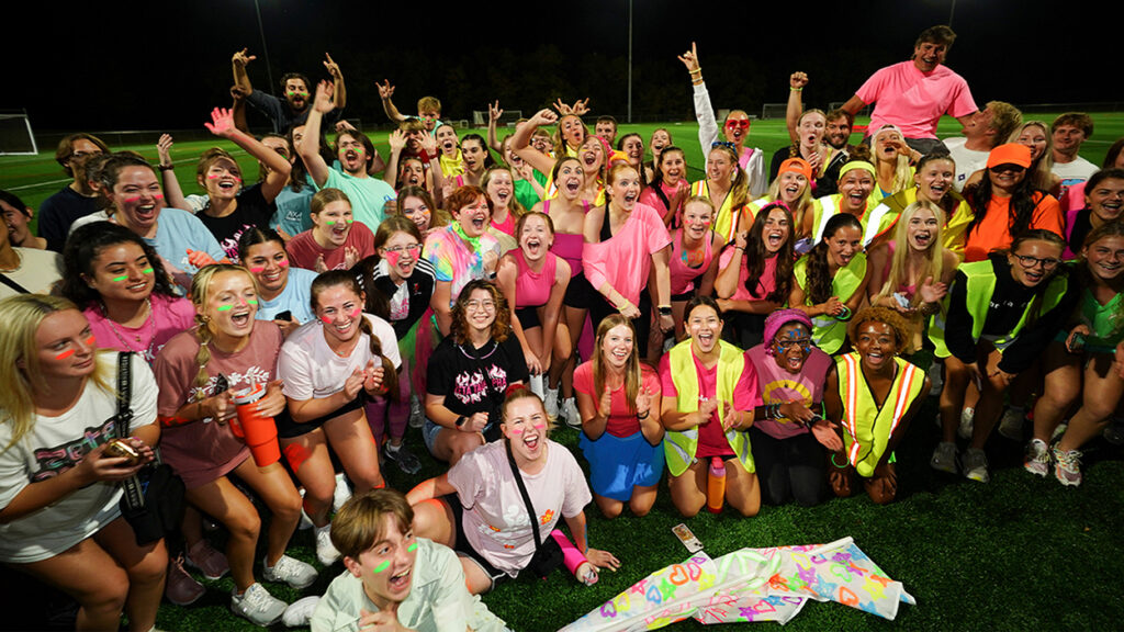 A large group of students wearing bright neon colors and face paint cheering together on an athletic field at night during Greene Games.