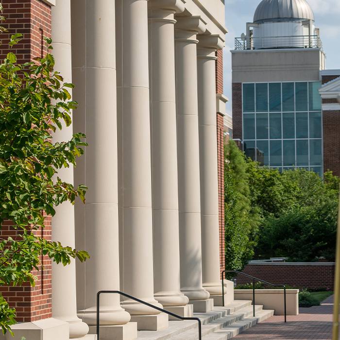 Exterior view of white stone columns alongside the Pryor Learning Center with the campus observatory in the background.