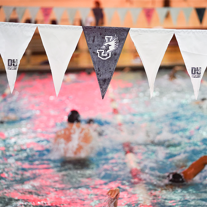 Swimmers in motion in a pool framed in the foreground by pennant flags, including 1 black flag featuring the William Jewell College Cardinal logo.