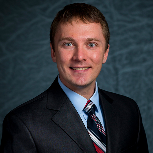 Headshot of Dr. Yuriy Bots, Professor of Economics and Business Administration at William Jewell College, wearing a dark suit jacket, blue shirt, and red and silver striped tie against a textured gray backdrop.