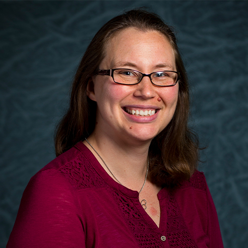 Headshot of Dr. Maggie Sherer, Professor of Physics at William Jewell College, wearing glasses and a deep red lace-trimmed top against a textured gray backdrop.