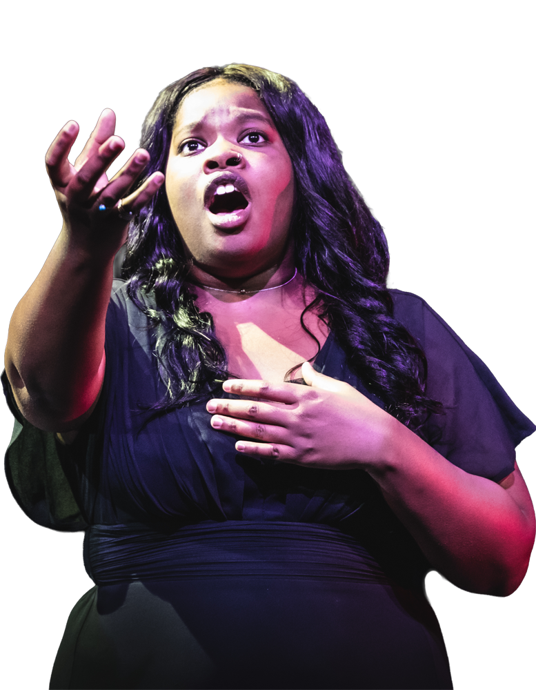 Carlyn Schrock singing on stage with hand outstretched under purple performance lighting.