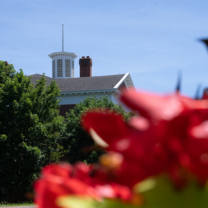 The white cupola of Jewell Hall rising above lush green trees, framed by out-of-focus red flowers in the foreground.