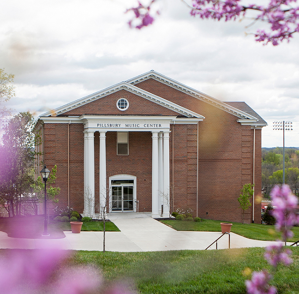 An exterior view of Pillsbury Music Center with white columns framed by purple spring blossoms.