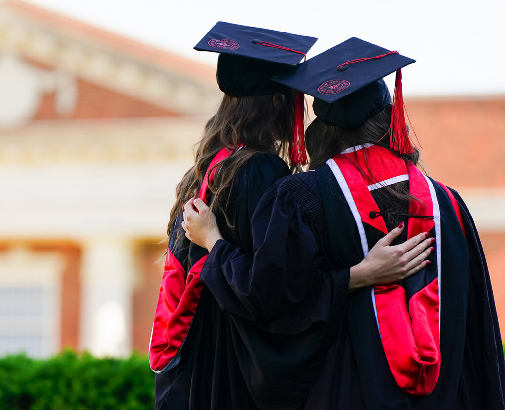 2 graduates in black gowns, red hoods, and mortarboards embracing while looking toward campus.