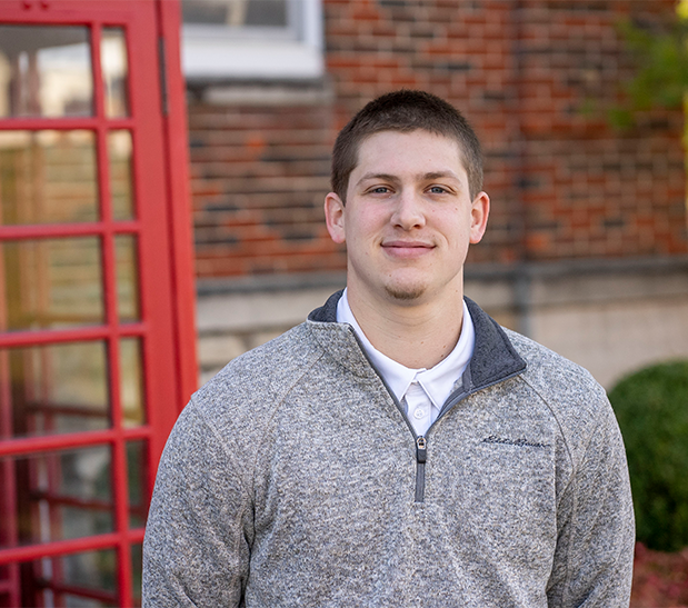 Portrait of John Zimmerman, wearing a light gray quarter-zip sweater in front of a red telephone booth.