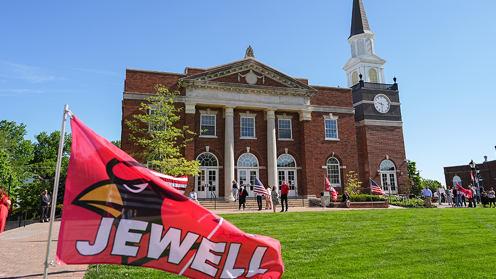 A red William Jewell flag waving on the lawn in front of Gano Chapel.