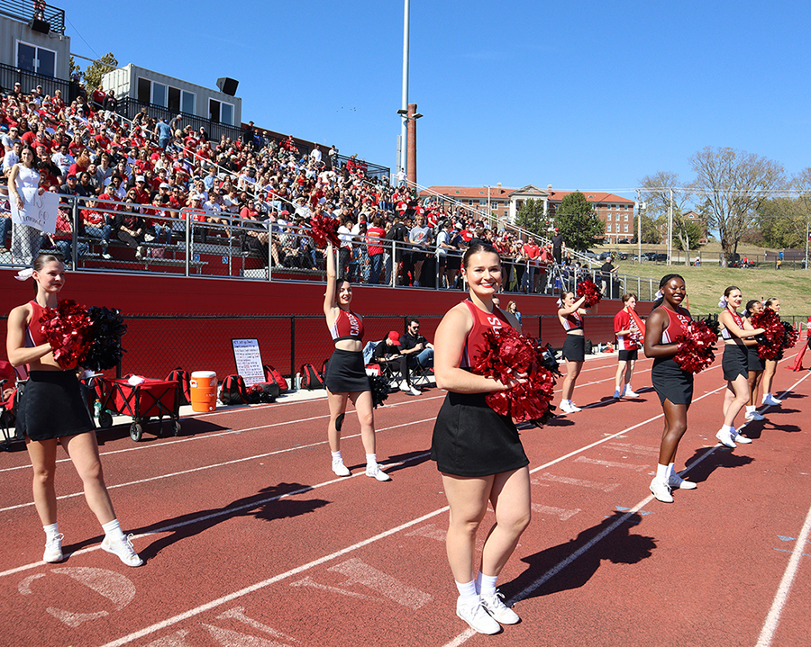 William Jewell cheerleaders lined up on the sideline track performing with pom-poms in front of filled stadium bleachers.