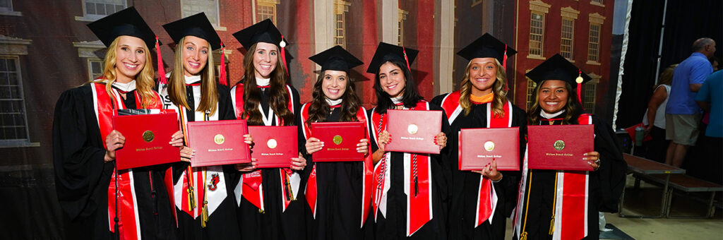 7 female graduates in black caps and gowns holding red diploma covers at commencement.