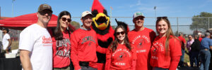 6 alumni and fans in William Jewell red apparel posing with Billy J. Bird outdoors at a homecoming event.