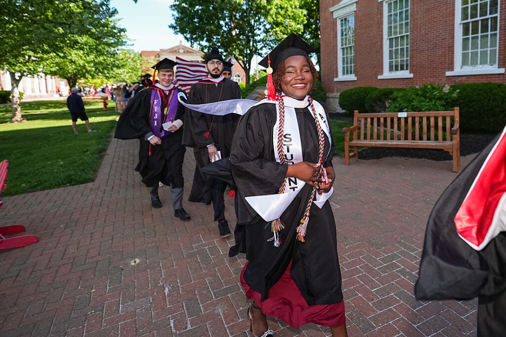 Tatyana Turner smiling in a black cap, gown, and white stole in a procession of graduates along a brick path.