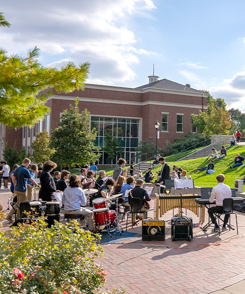 The William Jewell College jazz band performing outdoors on a brick quad with students sitting on the lawn in front of campus buildings.