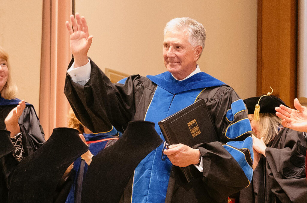 President Drew Van Horn waving to an applauding crowd while wearing academic regalia at a campus ceremony.