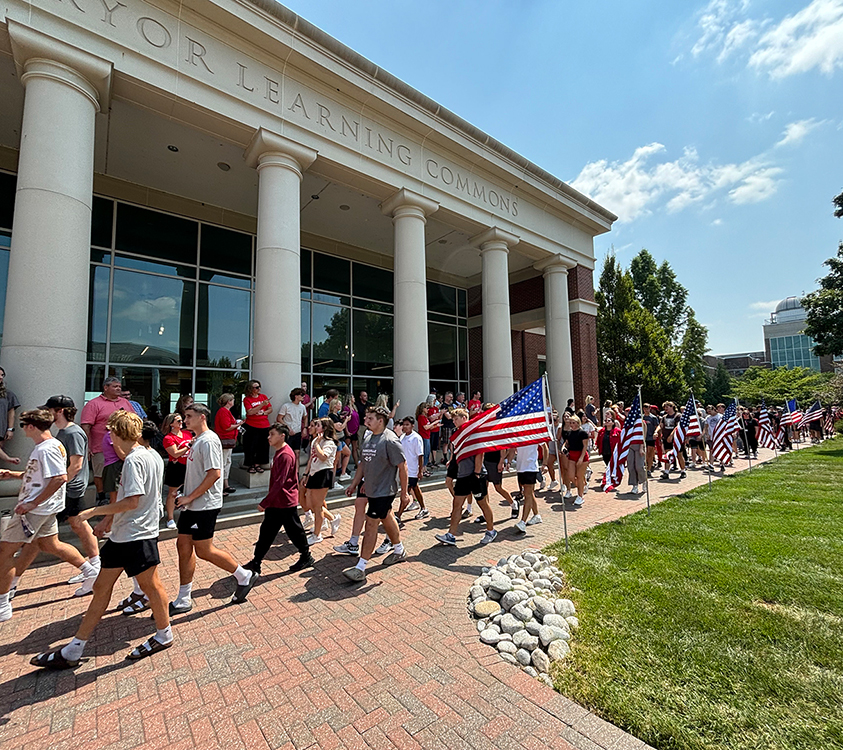 Students walking along a brick pathway outside Pryor Learning Commons during an orientation event while carrying American flags.