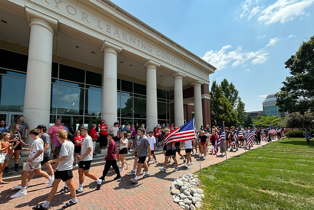A crowd of students walking along a brick walkway past Pryor Learning Commons holding American flags during campus orientation.