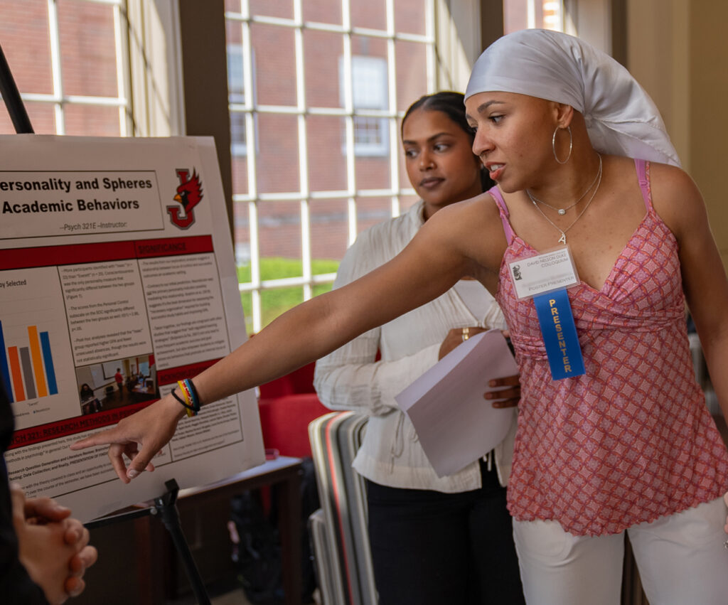 A female student wearing a white headscarf presenting a psychology research poster to an attendee.