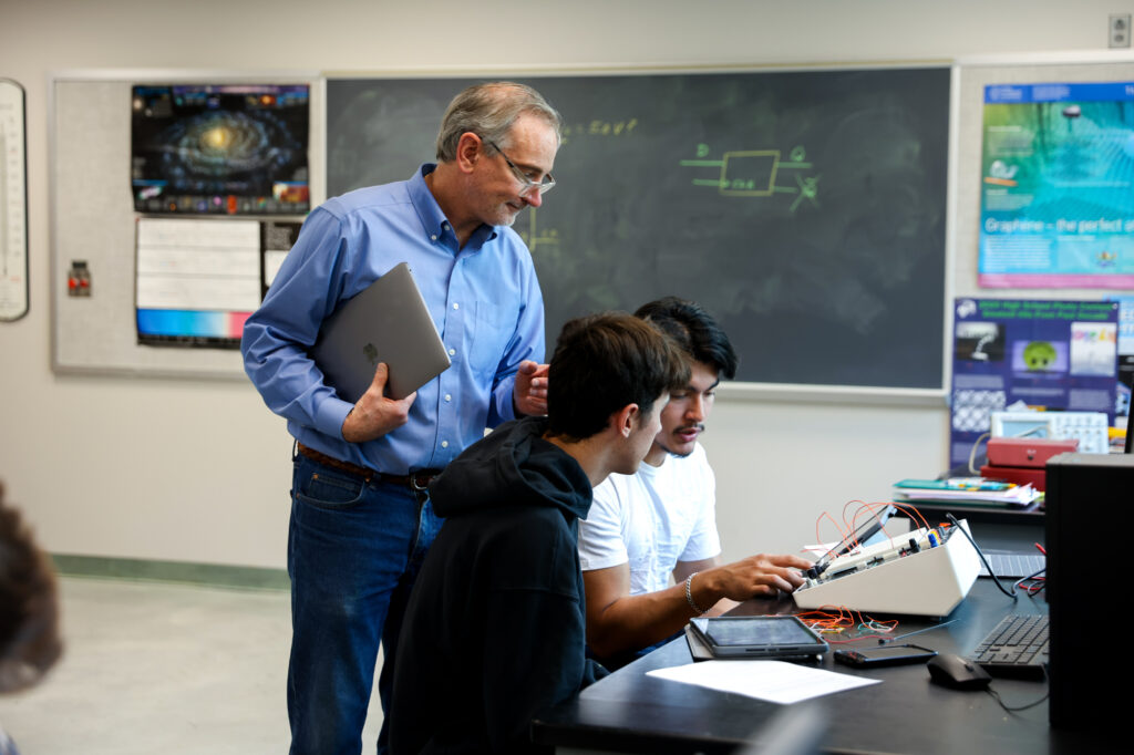 A male professor holding a laptop and assisting 2 male students working on an electronics lab project.