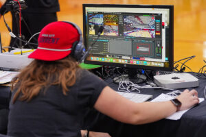 A student wearing headphones and a red Kansas City Current hat operating a live video broadcast interface on a desktop computer.