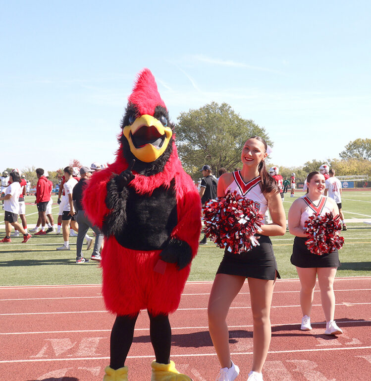 A cheerleader holding red and black pom-poms posing beside Billy J. Bird on the sideline track.