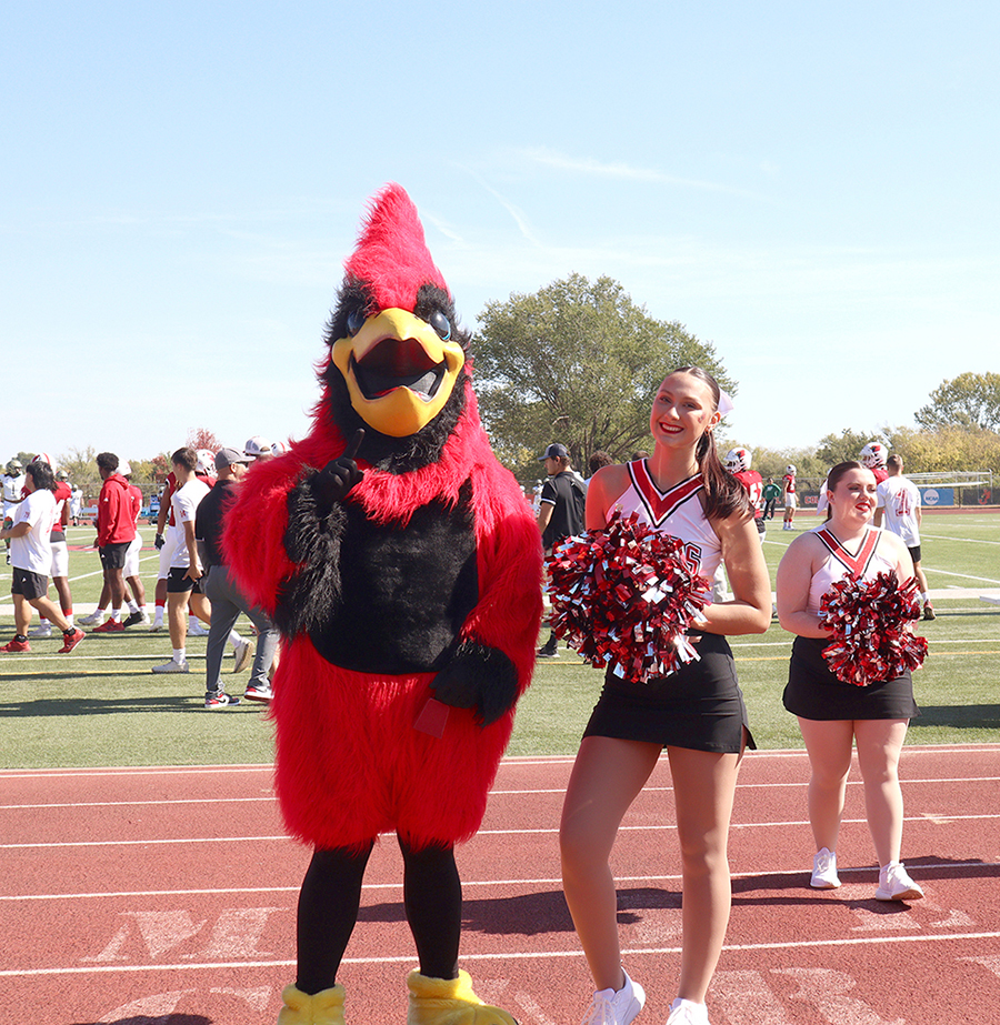 A cheerleader holding red and black pom-poms posing beside Billy J. Bird on the sideline track.