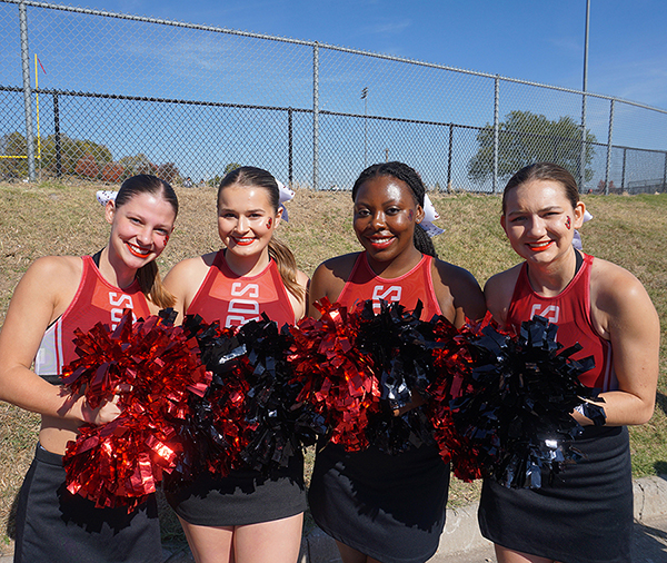 4 William Jewell cheerleaders holding red and black pom-poms while smiling together.