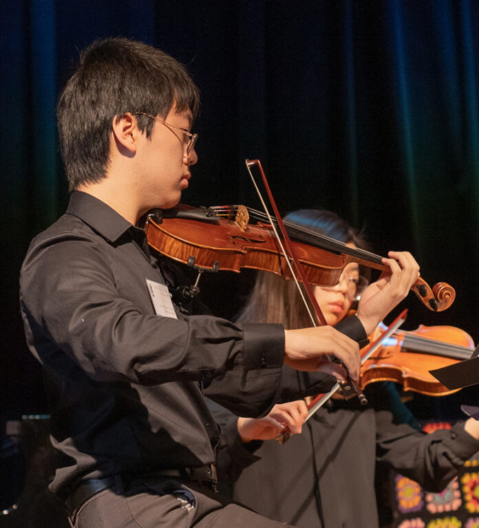 A male student in a black button-down shirt playing the violin during a musical performance.