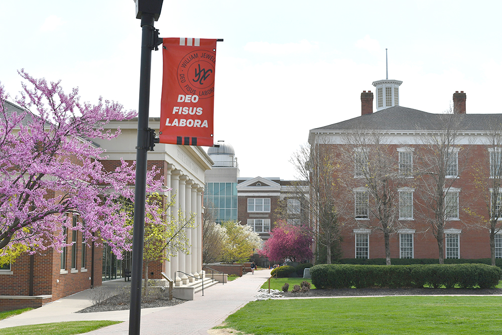 A lamp post banner reading 'Deo Fisus Labora' in front of Pryor Learning Commons during spring.
