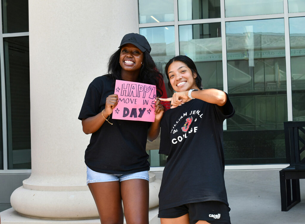 2 female students smiling and holding a handmade 'Happy Move In Day' sign outside a campus building.