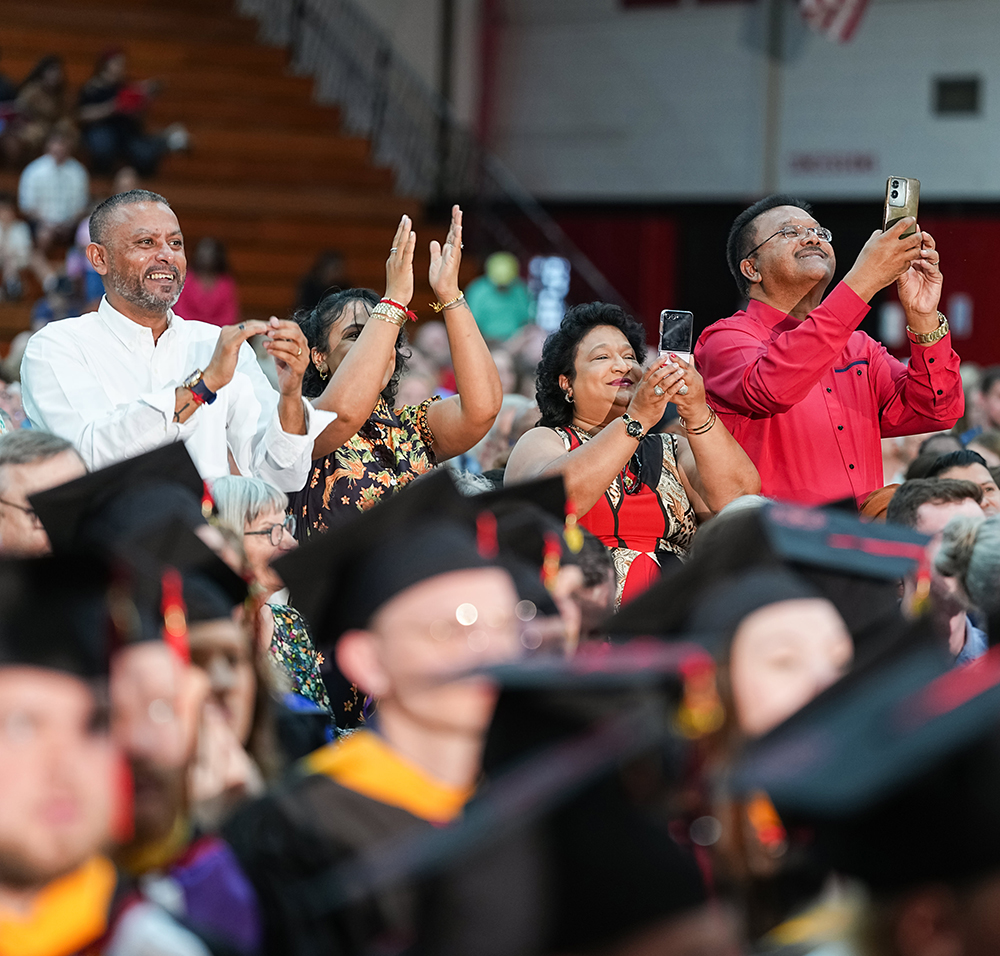 Family members applauding and taking photos on mobile phones during a graduation ceremony behind seated graduates in caps and gowns.