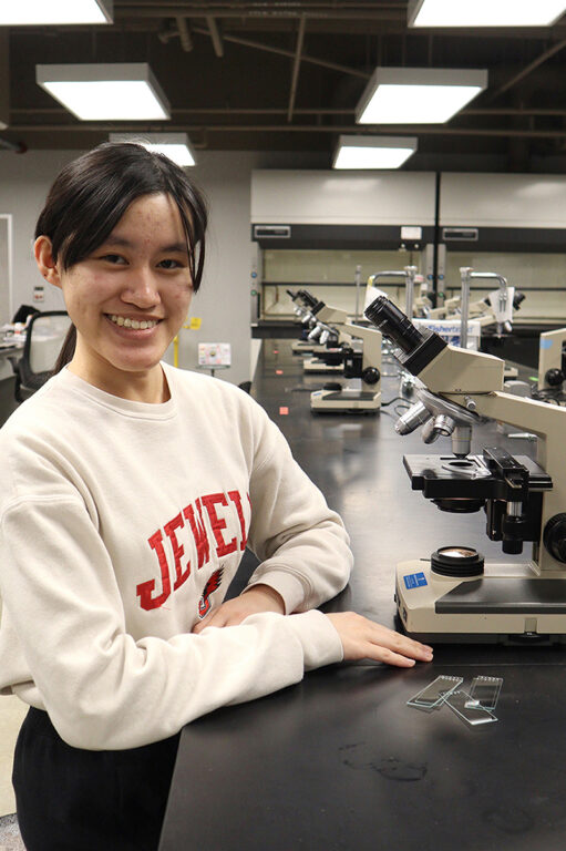 A female student in a Jewell sweatshirt smiling beside a lab microscope with glass slides.