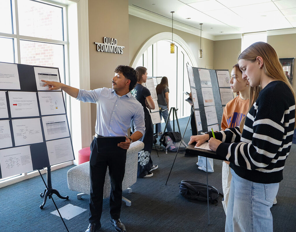 A male student presenting a research poster to 2 female students in Pryor Learning Commons.