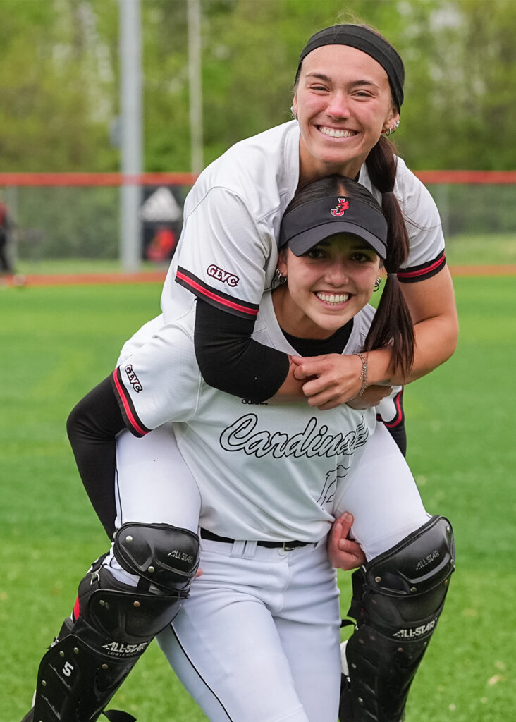 A William Jewell softball player smiling while carrying a teammate wearing catcher's gear on her back.