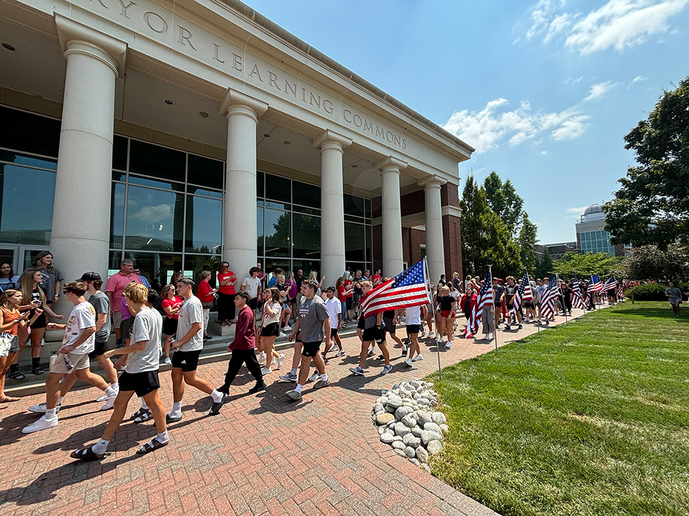 Students carrying American flags and walking in a procession past Pryor Learning Commons.