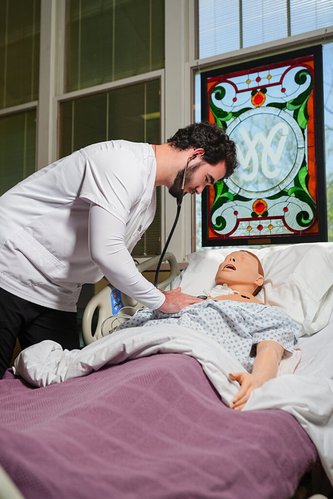 A nursing student using a stethoscope on a medical training mannequin in a clinical simulation lab.