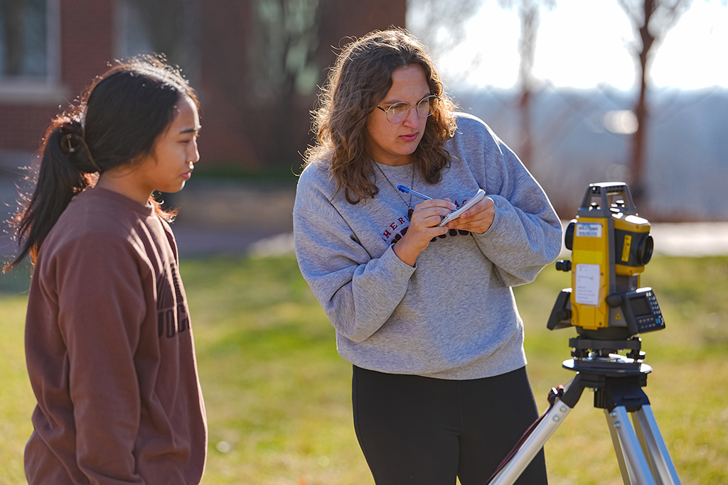 2 civil engineering students using a land surveying total station on campus while taking field notes.