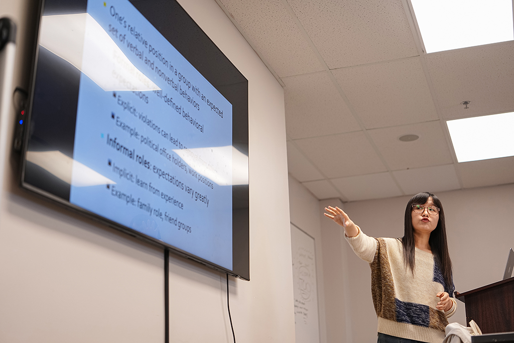 Ge Zhu, Assistant Professor of Communication, gesturing toward a digital presentation screen while teaching a classroom lecture.