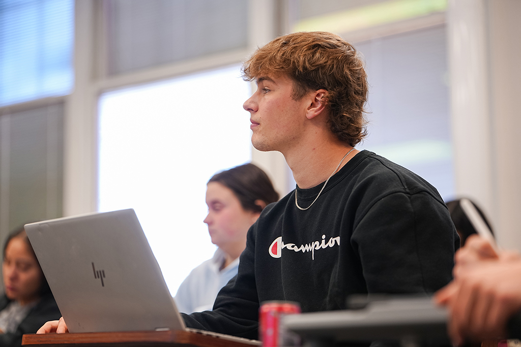A student sitting at a desk and taking notes on a laptop during a communication class lecture.
