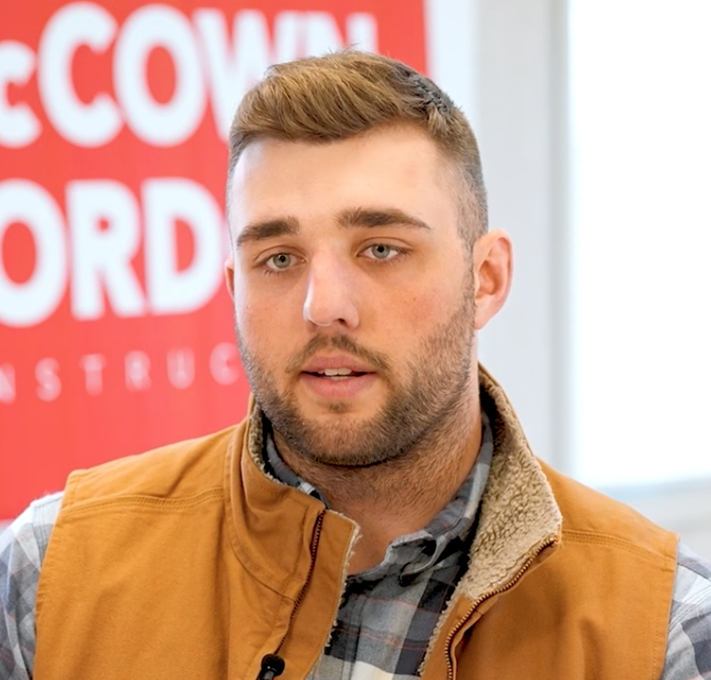 Major Walker, a civil engineering alumnus and field engineer wearing a tan vest and flannel shirt, speaking in front of a McCownGordon Construction banner.
