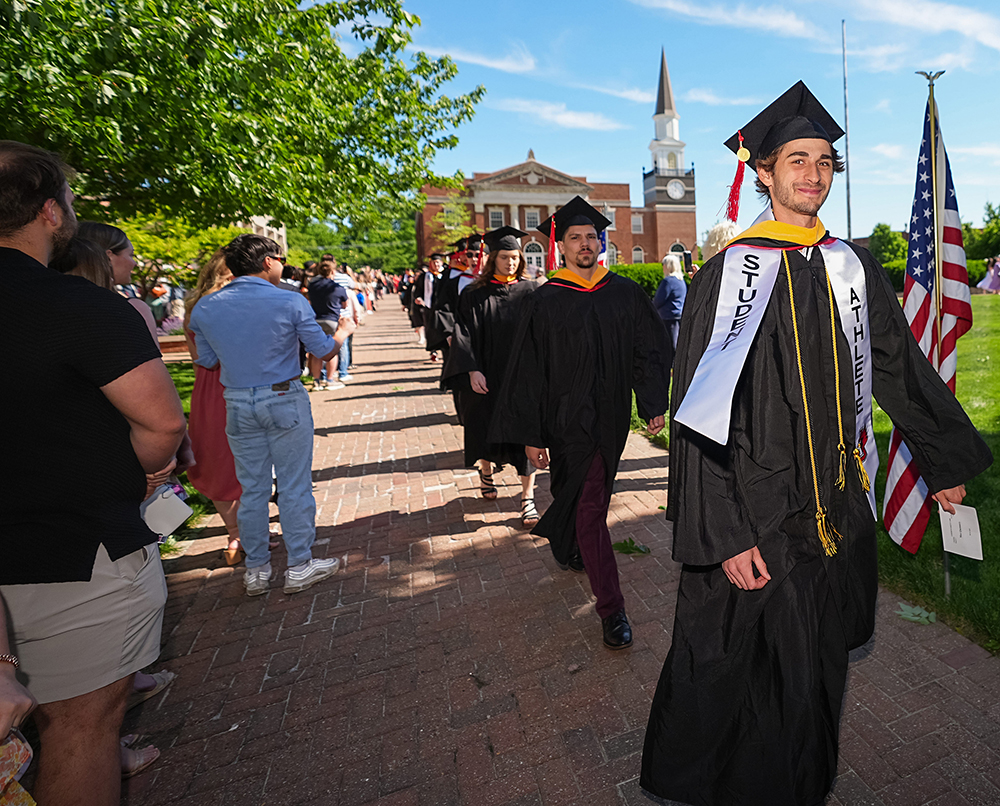 A graduating student-athlete smiling as he leads a procession of graduates along the brick walkway on the Quad.