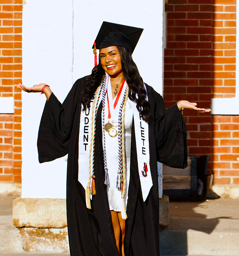 Hrithika Bhugvandeen smiling in a black graduation cap and gown, wearing a white "Student Athlete" stole and multiple honor cords outside a brick campus building.