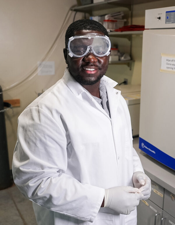 Derrick Ampofo, a biology student at William Jewell College, wearing a white lab coat, safety goggles, and protective gloves in a research laboratory.