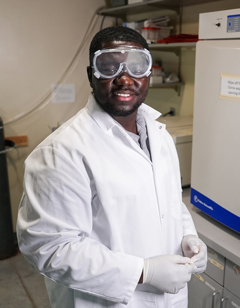 Derrick Ampofo, a biology student at William Jewell College, wearing a white lab coat, safety goggles, and protective gloves in a research laboratory.