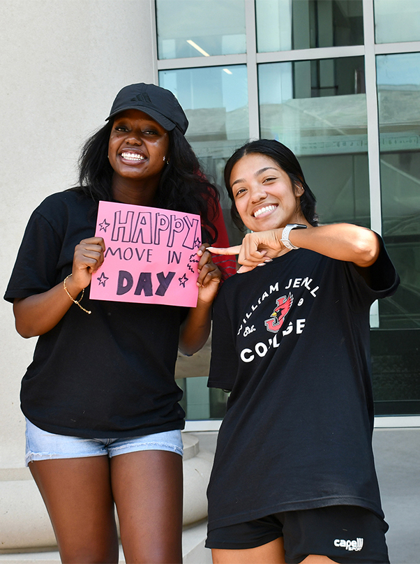 2 smiling students celebrating Move-In Day, with one holding a pink "Happy Move In Day" sign and the other pointing toward it.
