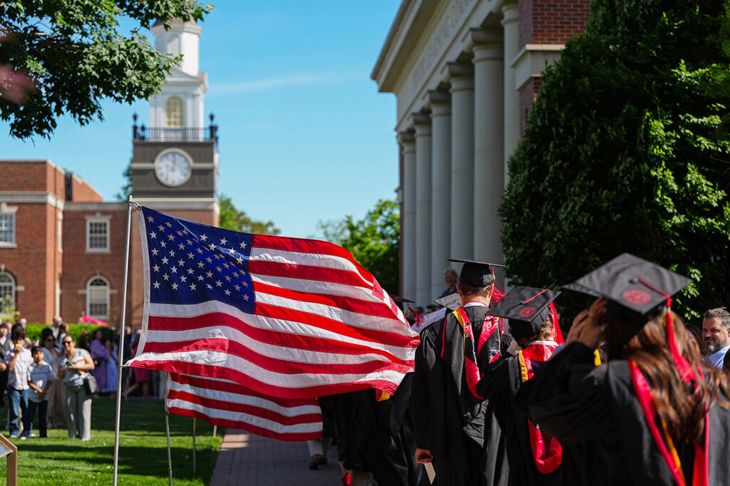 Graduating students in caps and gowns walking along the brick Quad pathway next to an American flag.