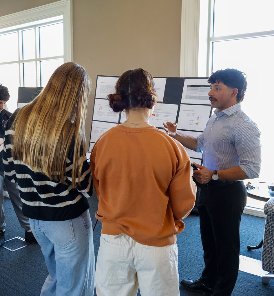 A male student presenting his research poster to two fellow students during a chemistry presentation in the Pryor Learning Commons.